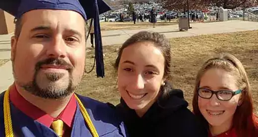 Daniel Stokoe wearing commencement gown with daughters Kayla in black hoodie and Kira in red.