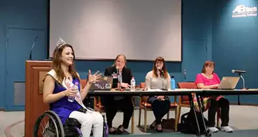 Madeline Delp in a wheelchair in front of three women at a table. 