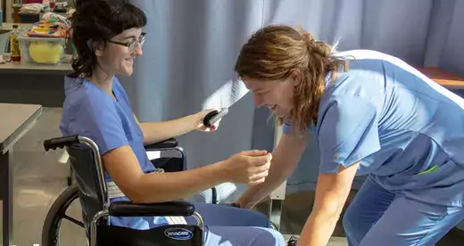 Two Nurse aide students. One assisting the other in a wheelchair
