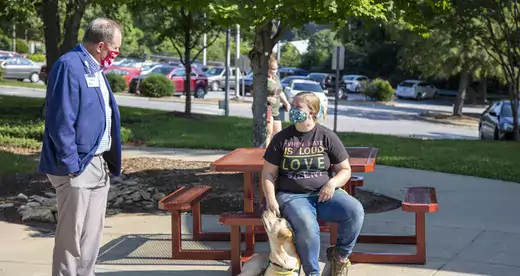 John Gossett standing in front of a student at a picnic table with dog sitting at student's feet. 