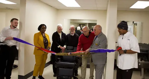 Seven people in front of a barber chair cutting a ribbon