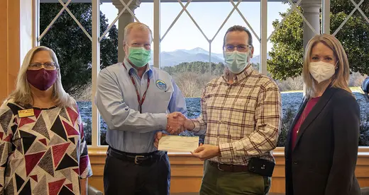 Four people standing by a window with two men exchanging a check