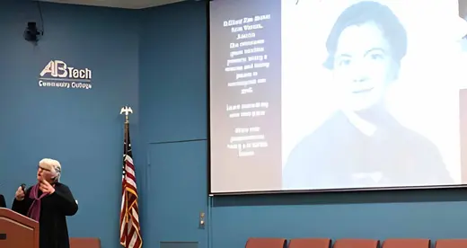 Woman at a lectern with a woman on the screen behind her. 