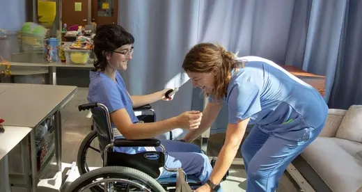 Two Nurse aide students. One assisting the other in a wheelchair