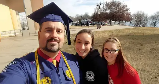 Daniel Stokoe wearing commencement gown with daughters Kayla in black hoodie and Kira in red.
