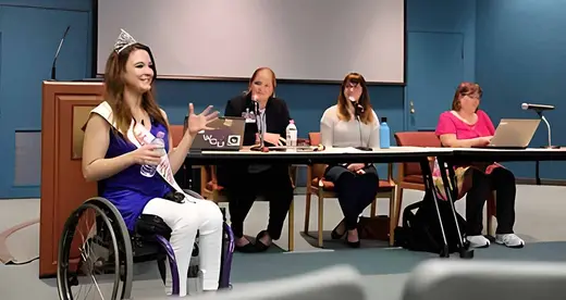 Madeline Delp in a wheelchair in front of three women at a table. 