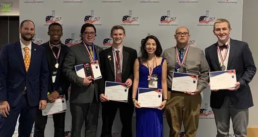 Seven well dressed chefs standing in front of a wall holding certficates