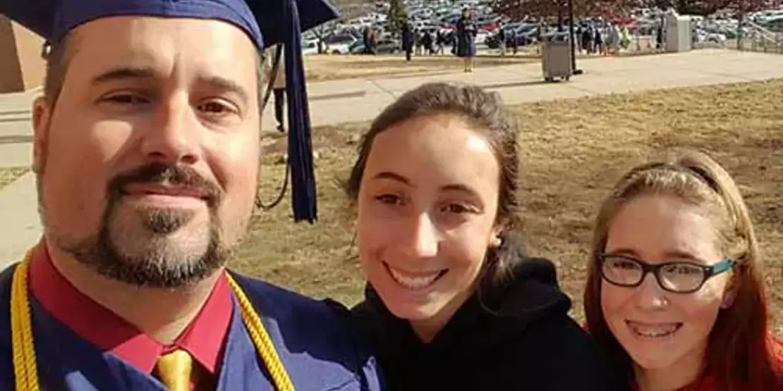 Daniel Stokoe wearing commencement gown with daughters Kayla in black hoodie and Kira in red.