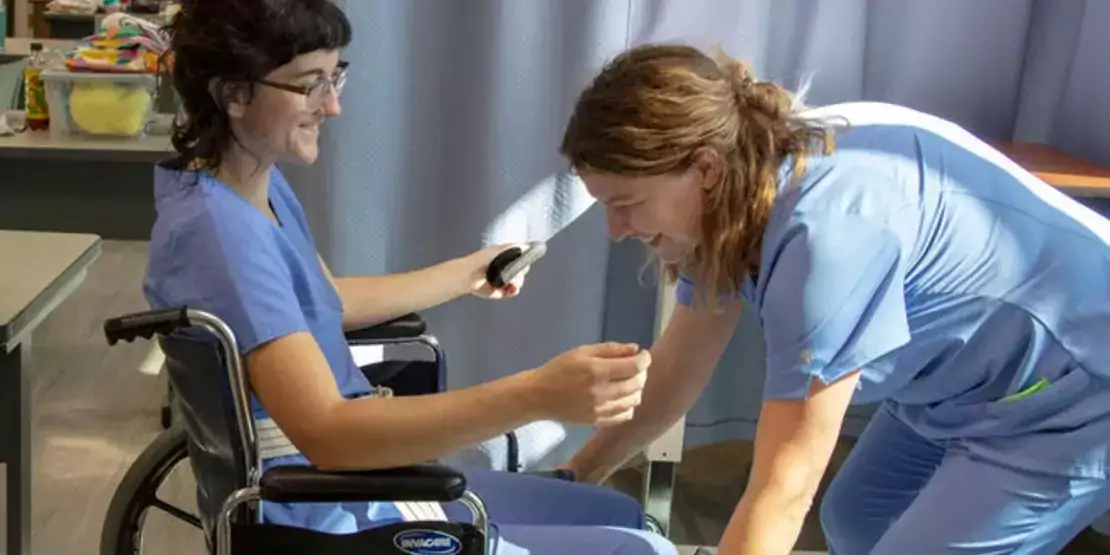 Two Nurse aide students. One assisting the other in a wheelchair