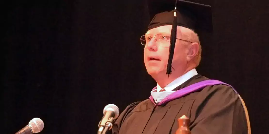 Max Queen wearing academic regalia and speaking at lectern