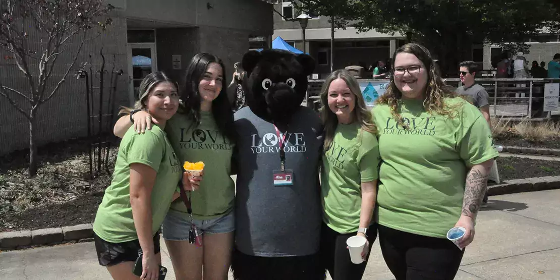 2 A-B Tech students posing with the A-B Tech Trailblazer bear wearing lime green Earth Day t-shirts.