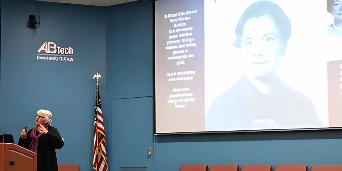 Woman at a lectern with a woman on the screen behind her. 