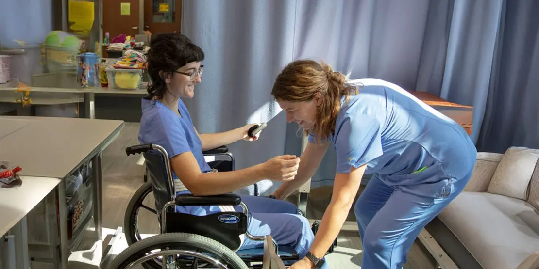 Two Nurse aide students. One assisting the other in a wheelchair