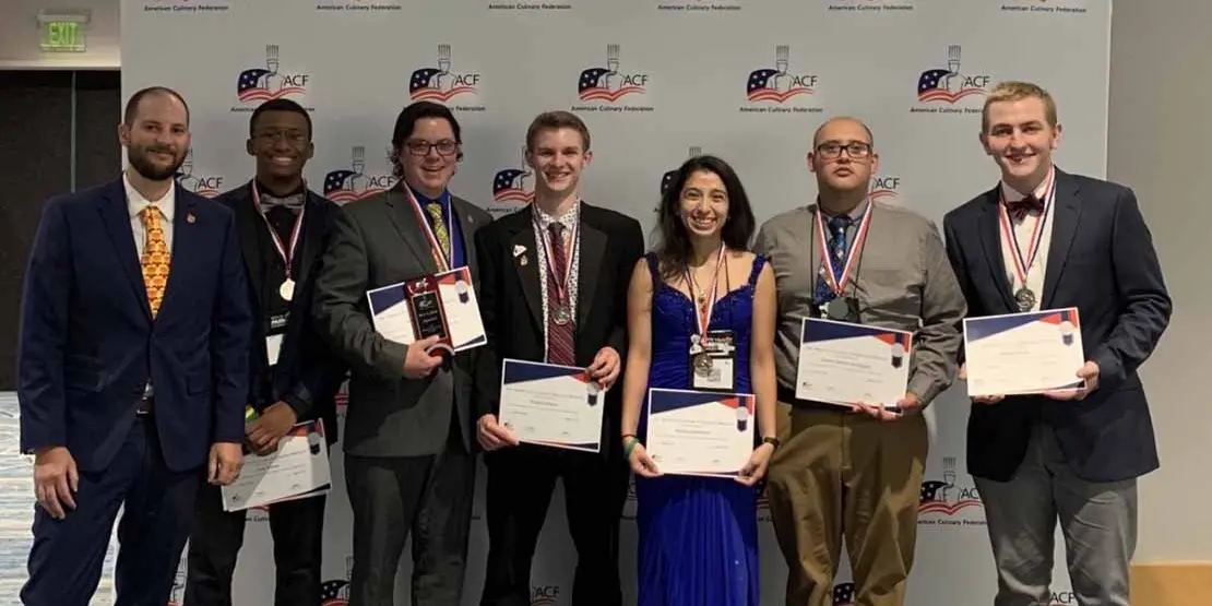 Seven well dressed chefs standing in front of a wall holding certficates