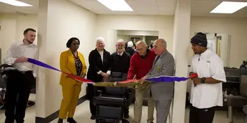 Seven people in front of a barber chair cutting a ribbon