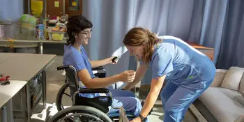 Two Nurse aide students. One assisting the other in a wheelchair